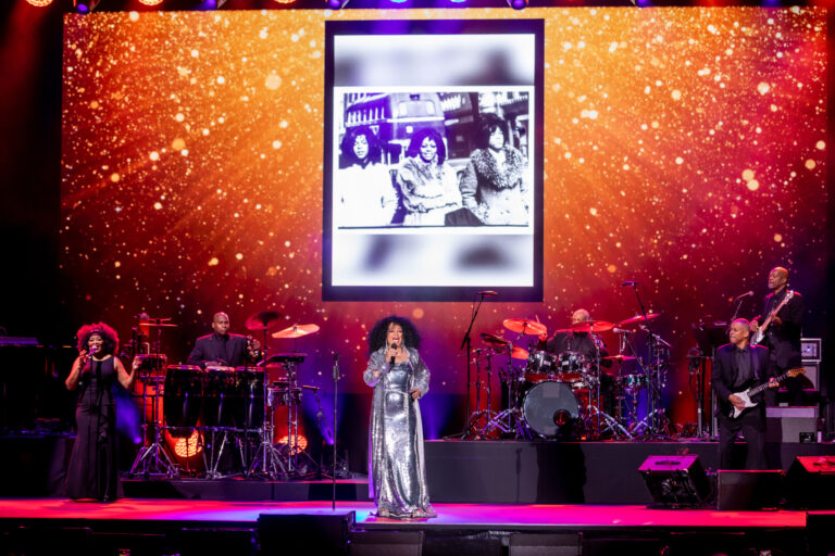 A singer performs onstage beneath a large illuminated screen showing a framed collage, with musicians across the stage.