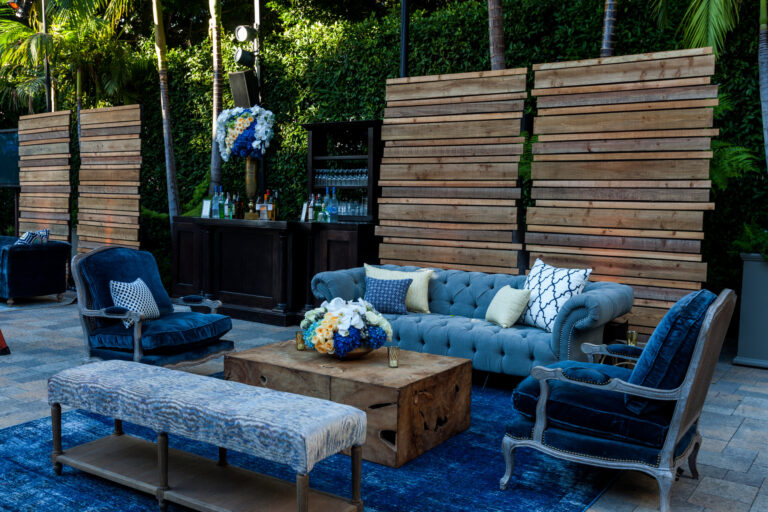 Lounge seating, low tables and a blue rug are arranged in a shaded outdoor patio beneath string lights.