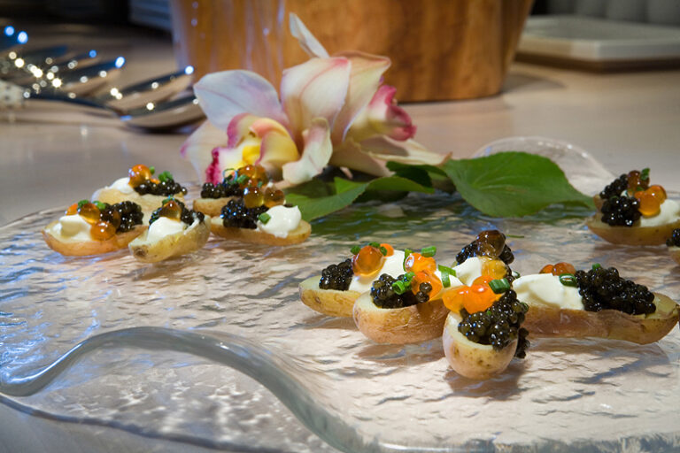A row of small plated appetizers topped with garnishes is arranged on a serving surface beside a flower.