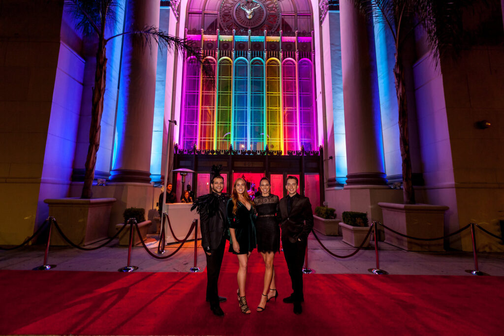 Four adults pose on a red carpet before a tall arched window lit in multiple colors.