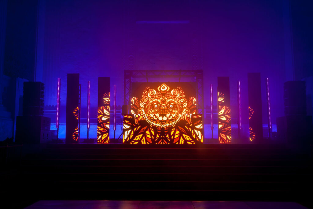 A glowing orange ornamental face fills the center of a purple-lit stage.