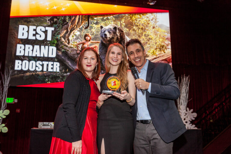 Three adults pose together in front of a screen reading Best Brand Booster, with a dog visible on the display.