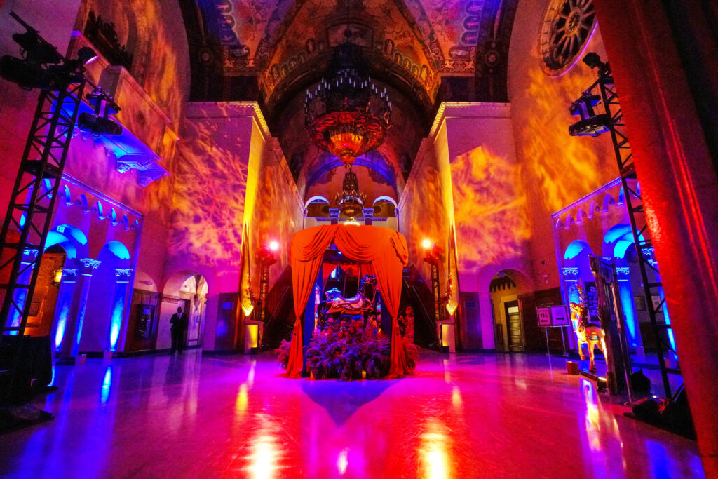 A grand arched hall is washed in orange and blue light around a red-draped carousel display.