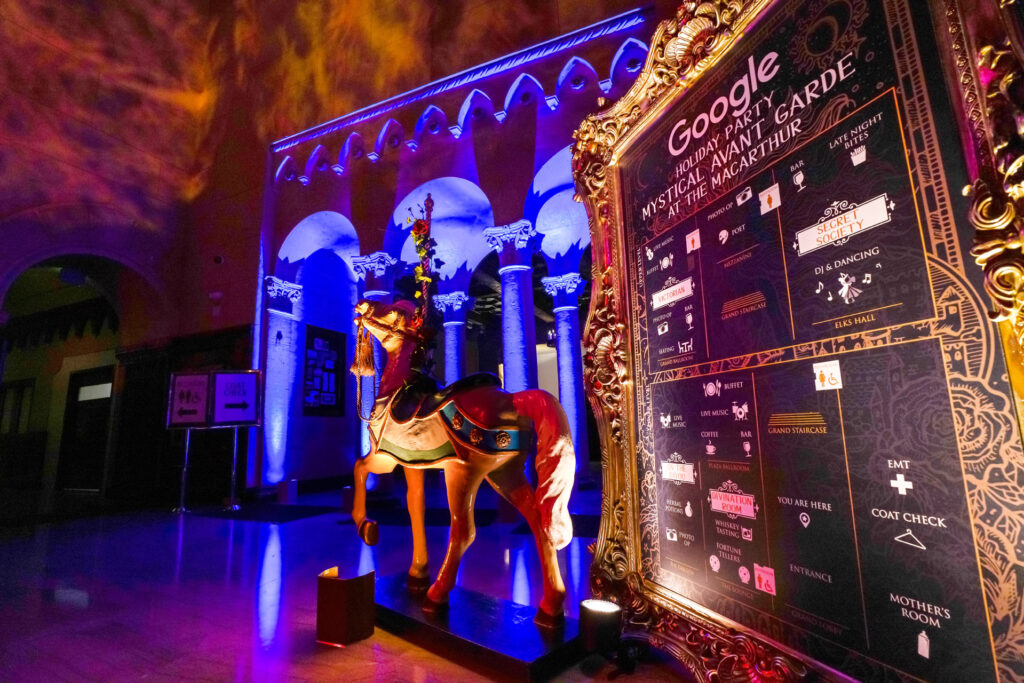 A gold carousel horse stands beside a Google Holiday Party wayfinding board in a blue-lit hall.
