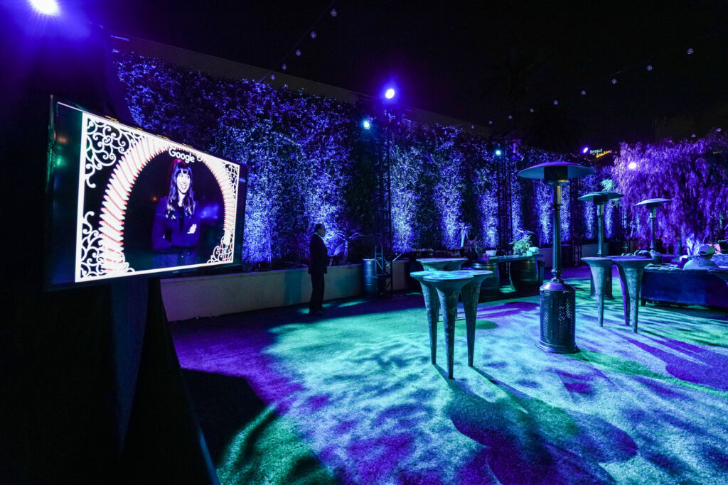 A large screen displays a portrait and Google logo beside cocktail tables in a blue-lit event space.