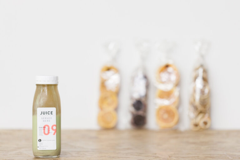 A small glass juice bottle stands in focus with three taller bottled drinks softly blurred behind it.
