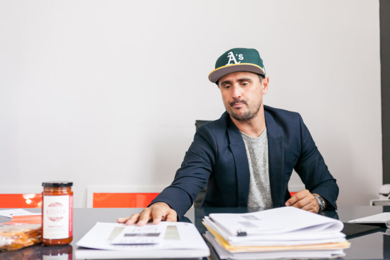 Man in a green cap and dark blazer seated at a desk with papers and juice products.