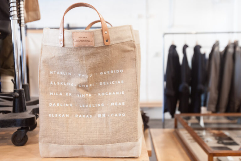 Tan canvas tote with leather handles and rows of multilingual lettering displayed beside clothing racks.