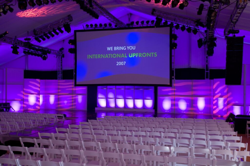 Rows of chairs facing a large presentation screen beneath purple event lighting.
