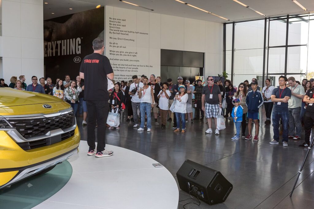 A presenter addressing attendees beside a yellow Kia display vehicle.